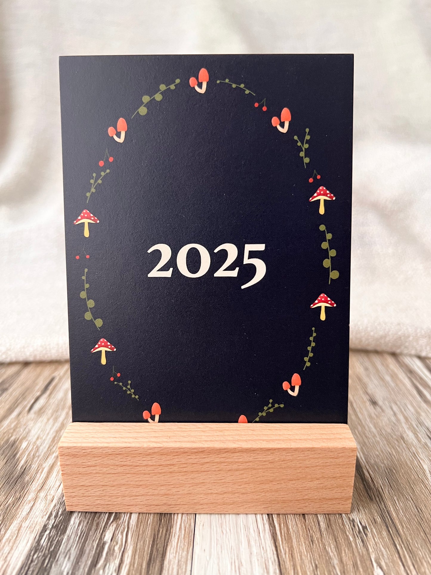 2025 desk calendar cover in post card size, inserted into wooden stand displayed on a wooden desk. 