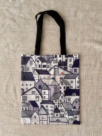 The image shows a cotton tote bag, with illustration of rows and rows of houses. The houses are all in dark blue and white. There is a cat in red walking along the houses. The colour contrast makes the cat stood out.