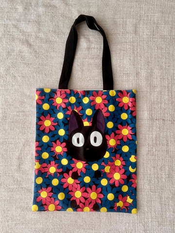 The image shows a colourful cotton tote bag. The illustration has a black cat with really big eyes hidden among pink and blue flowers. The colour contrast each other and makes the cat stood out. It's cute and adorable, perfect for a cat lover