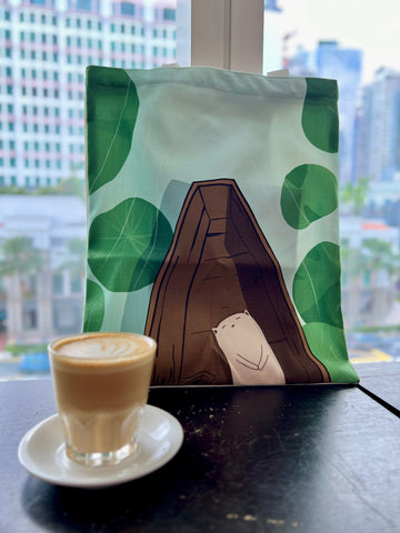 The image shows a cotton tote bag, with the illustration of a cat resting on a boat, floating along a lake full of lily pads. It is very relaxing, summer kind of vibe.