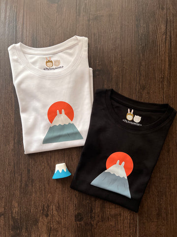 Fuji Bunny t-shirts in black and white, folded and placed on wood planks. The t-shirts featuring minimalist design of mount fuji with a pair of bunny ears and a rising orange sun in the background