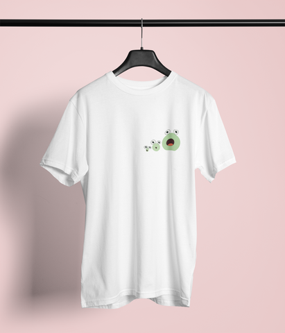 White t-shirt featuring a small, playful design of three green frogs, with their mouths wide opened on the upper left chest. The t-shirt hangs on a black hanger against a pinkish backdrop