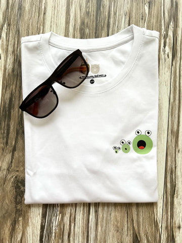 White t-shirt featuring a small, playful design of three green frogs, with their mouths wide opened on the upper left chest. The t-shirt is folded and placed on wooden plank.