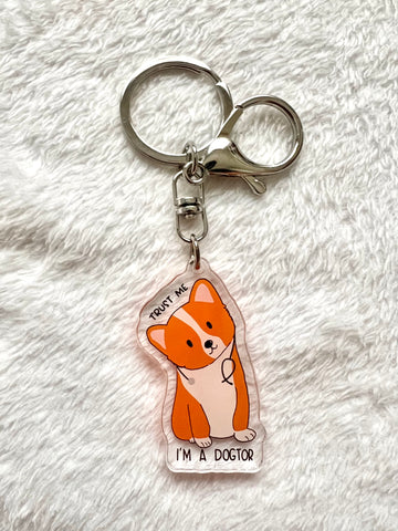 A corgi keychain lay flat on a furry mat. The corgi has its head tilted to one side, and had a stethoscope on its neck. There’s text written on it, “trust me, I’m a dogtor”. A play on the word doctor.