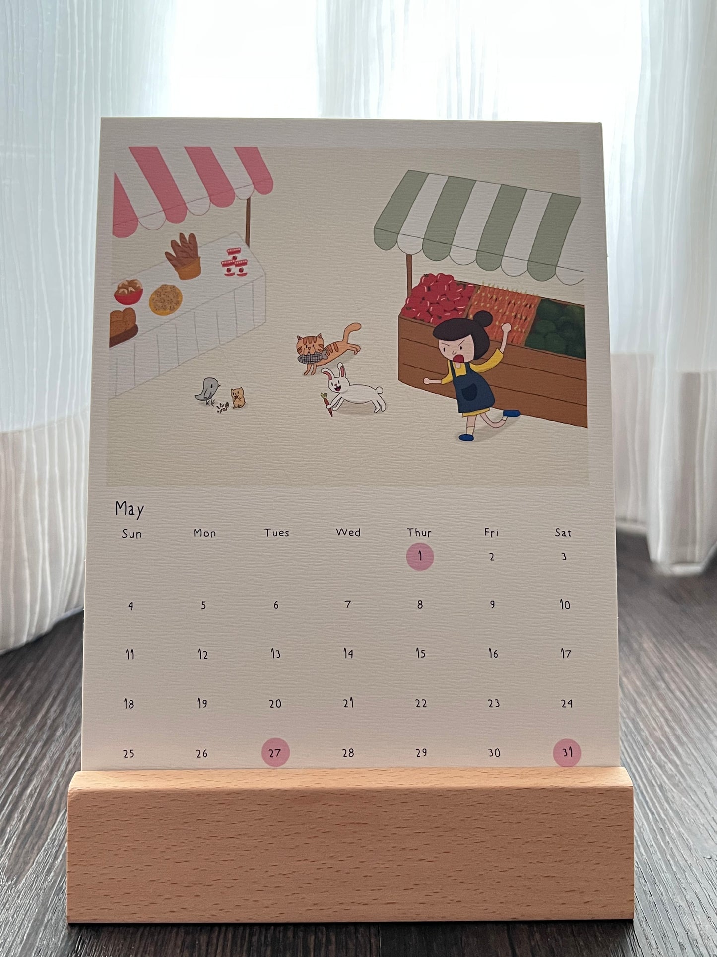 Calendar with the drawing of the cat, bunny. hamster and the pigeon getting food at a market. The Cat and The Bunny stole some food from the stall, hence got chased by an angry owner. The calendar is placed on a wooden stand