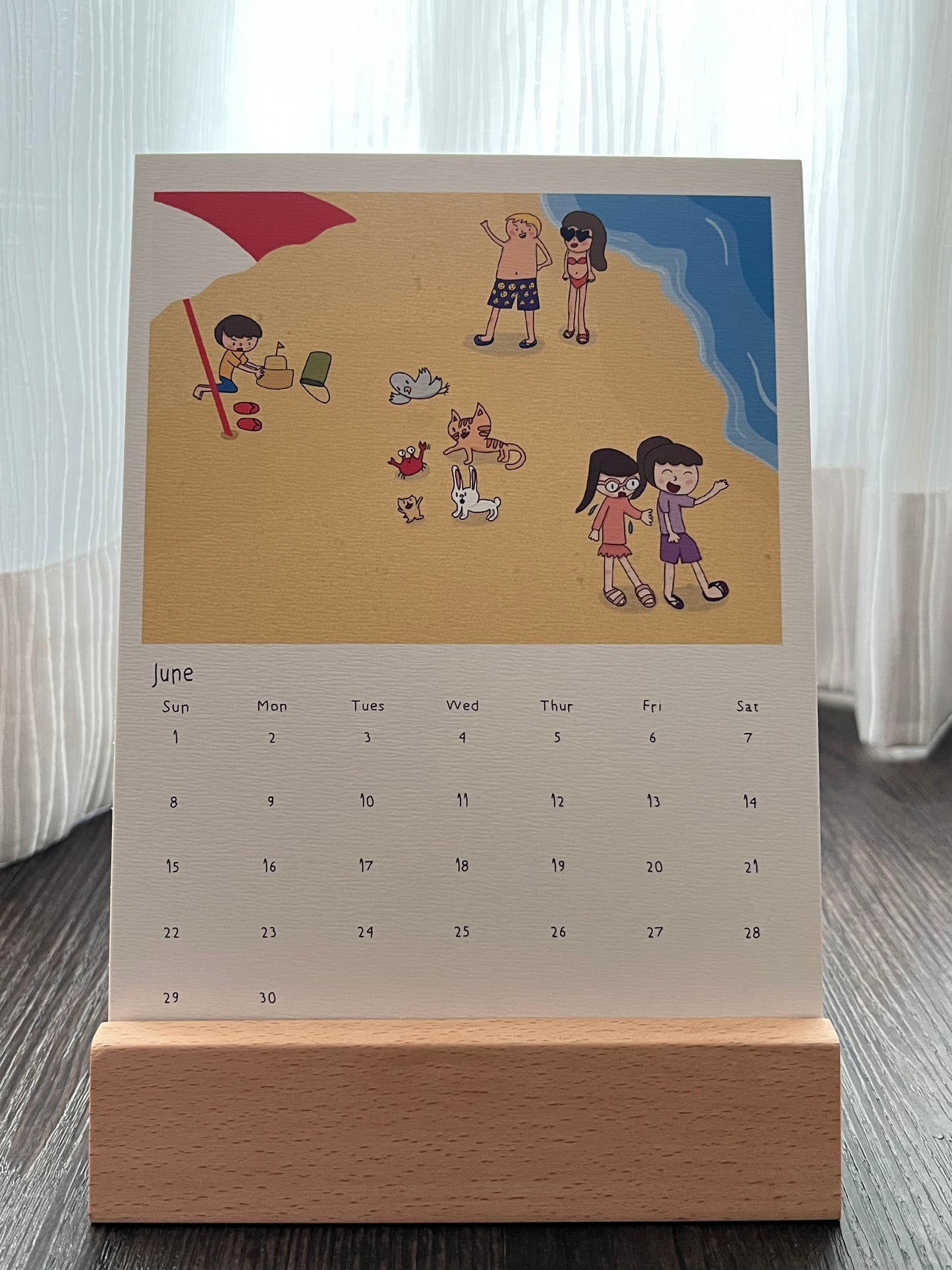 Calendar with the drawing of the cat, bunny, hamster and pigeon at the beach. They made friend with a crab. There were also some other beach goers at the beach. The calendar is placed on a wooden stand