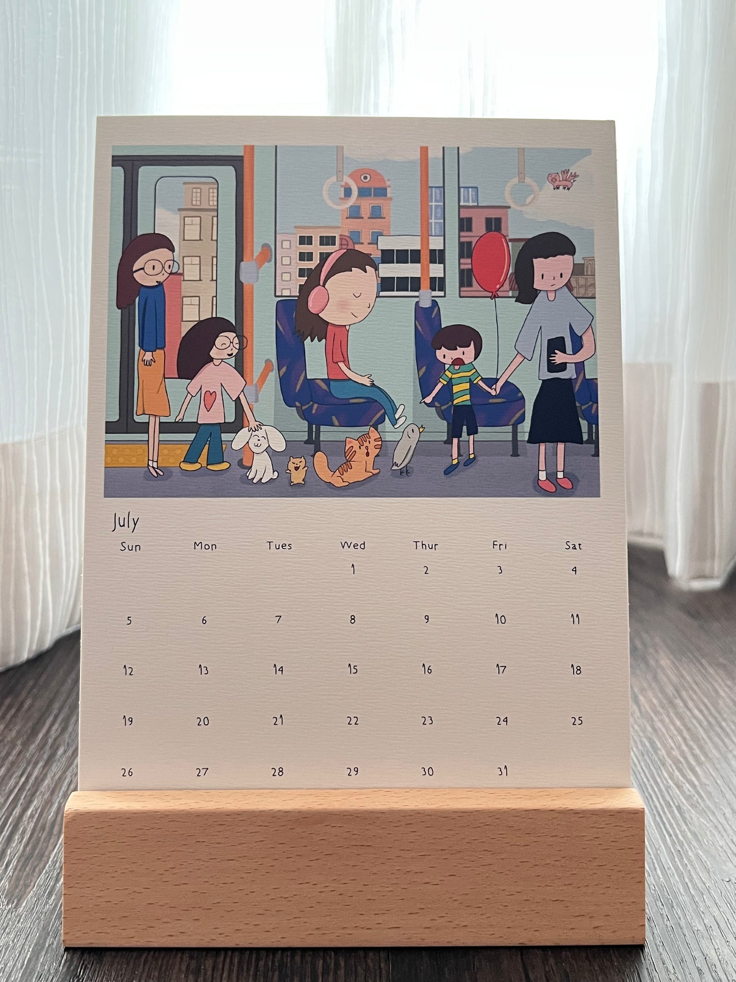 Calendar with the drawing of the cat, bunny, hamster and pigeon on a bus. The other passengers, especially the kids were amused by the scene. Some adults were just too engrossed with their screens. There's city scene outside the bus, and a flying pig. The calendar is placed on a wooden stand