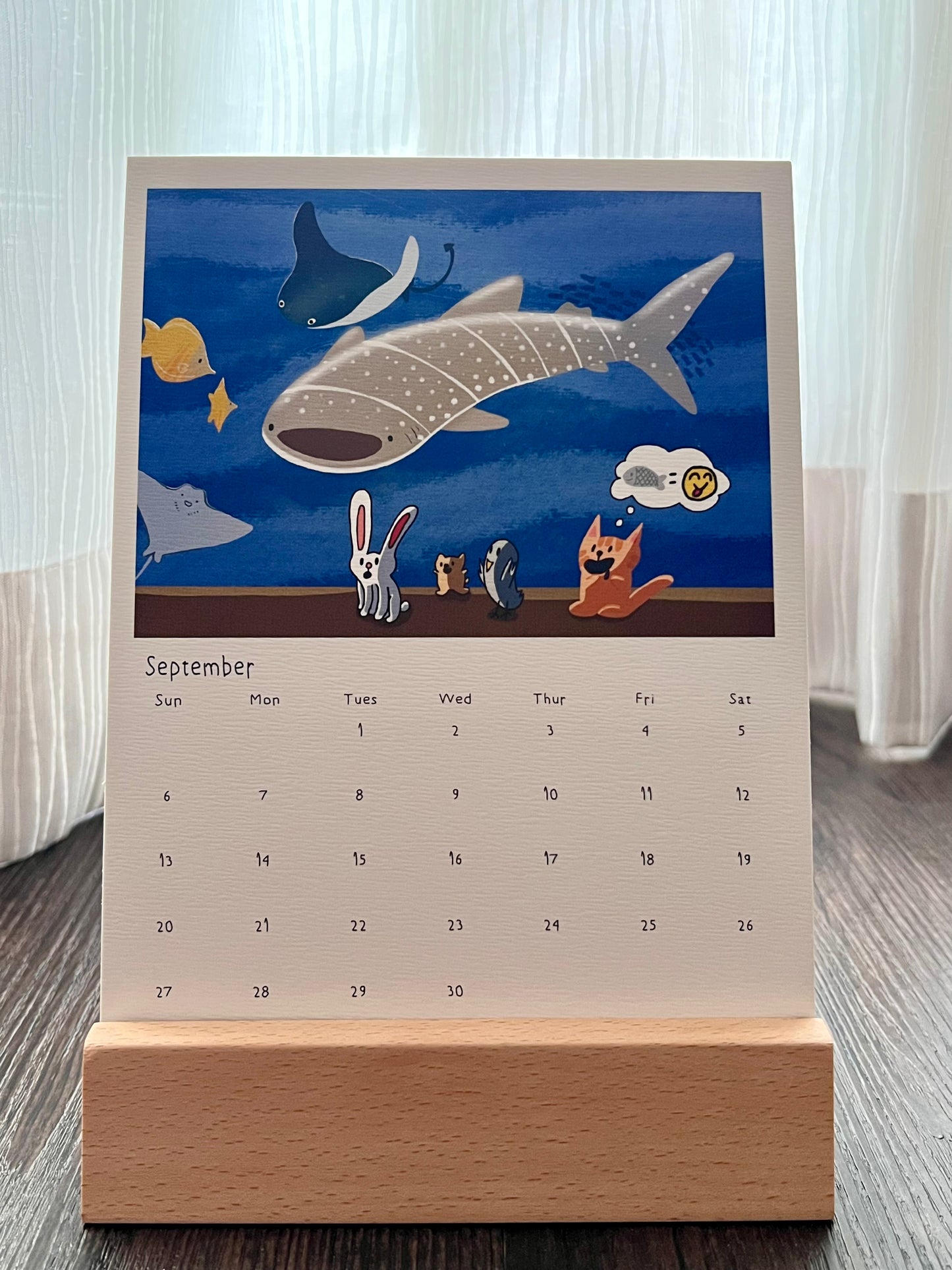 Calendar with the animal characters visiting aquarium. There was a whale shark, manta ray, sting ray in the huge tank. The animal characters are amused, and the cat is only thinking of food. The calendar is placed on wooden stand