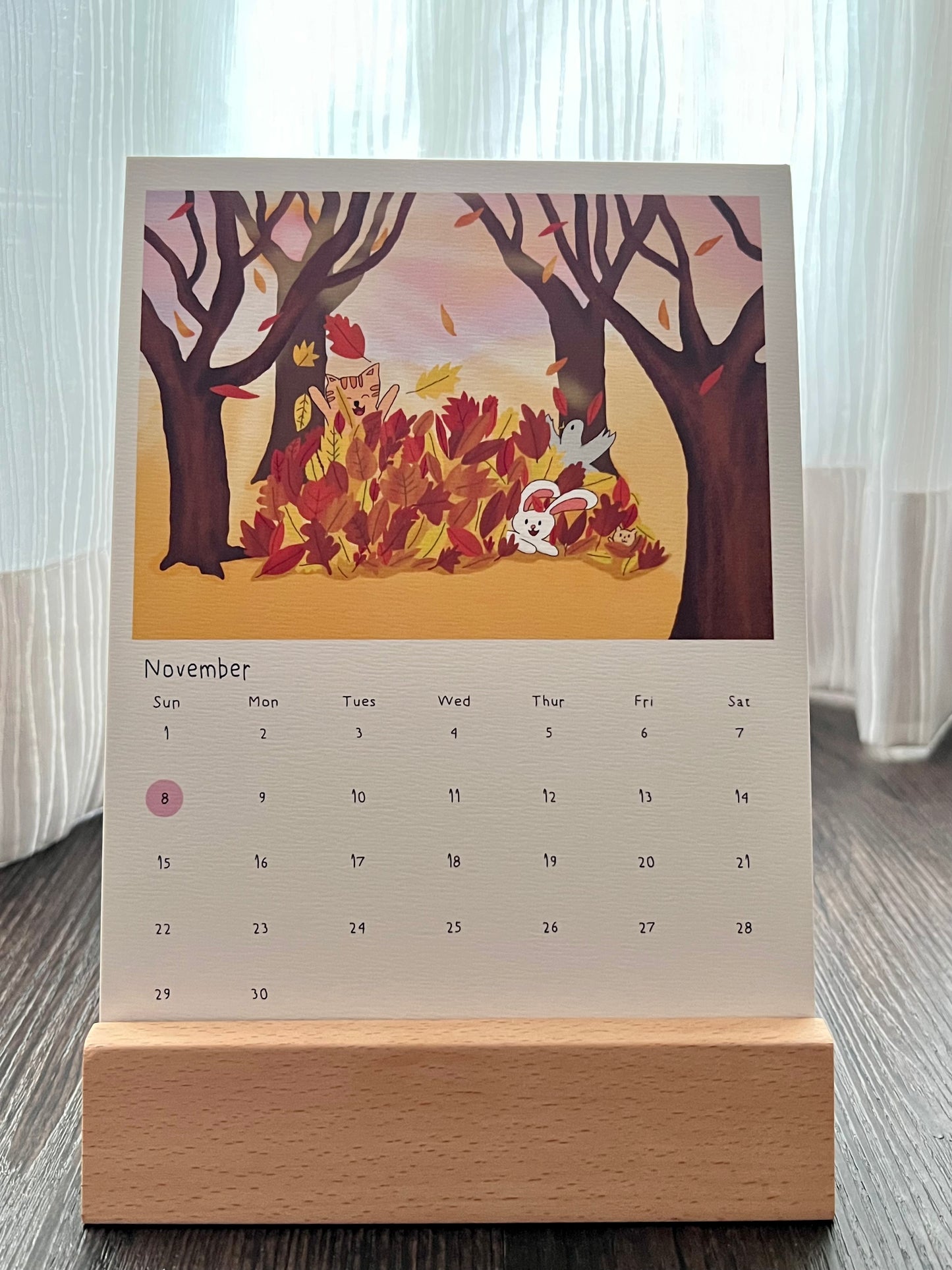 Calendar with autumn theme, the animal characters are playing with the fallen leaves. The calendar is placed on the wooden stand. Singapore public holiday is marked in pink