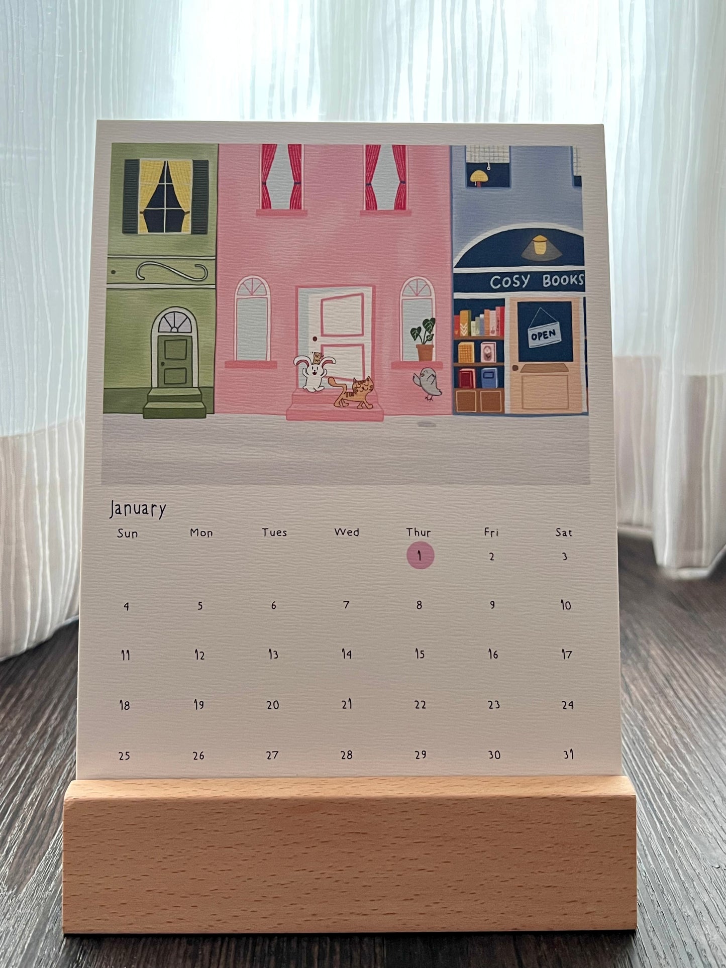 Calendar (January 2026) with a design of a street scene, where The Cat leading The Bunny and The Hamster to sneak out of their home. And they met their new friend, The Pigeon. Singapore Public Holiday is marked with pink circle. The calendar is placed on a wooden stand
