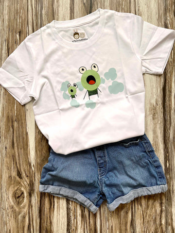 White T-shirt with a graphic design on the front. The design features two cartoonish, child drawn, wide-eyed frogs with expressive faces. The larger character, in the center, has a round face with a surprised expression, while the smaller character, to the left, has a similar expression. Both characters are surrounded by light blue, cloud-like shapes, adding a whimsical touch. The T-shirt is flat lay with a chilren's short against a wooden background