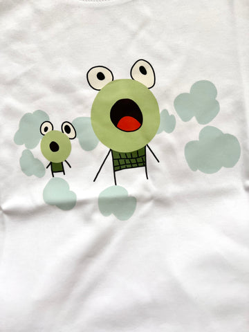 White T-shirt with a graphic design on the front. The design features two cartoonish, child drawn, wide-eyed frogs with expressive faces. The larger character, in the center, has a round face with a surprised expression, while the smaller character, to the left, has a similar expression. Both characters are surrounded by light blue, cloud-like shapes, adding a whimsical touch