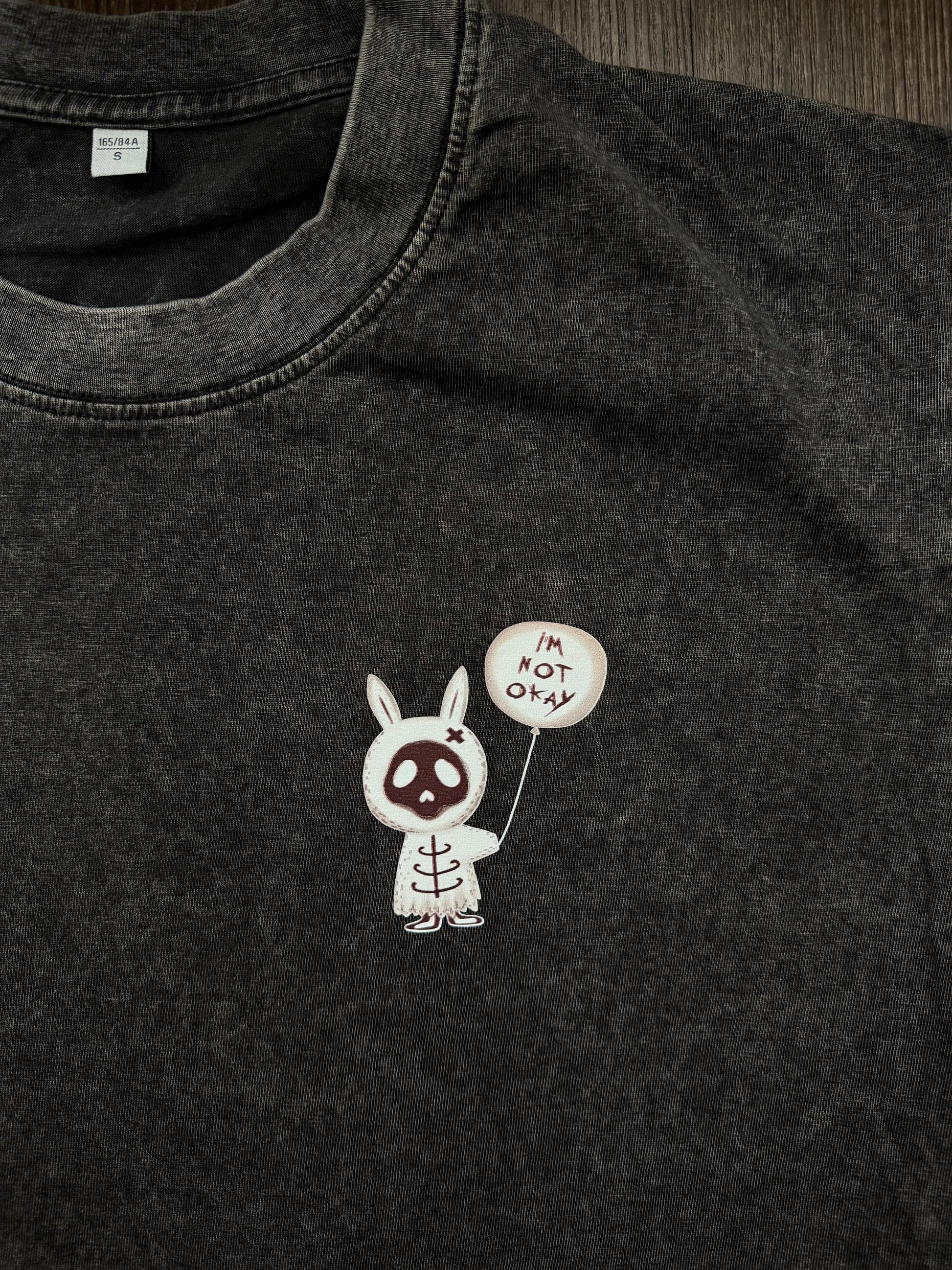 A close-up of a dark gray mineral-wash cotton t-shirt featuring a small graphic of a white bunny skeleton holding a balloon that says 'I'M NOT OKAY' in My Chemical Romance “The Black Parade” font