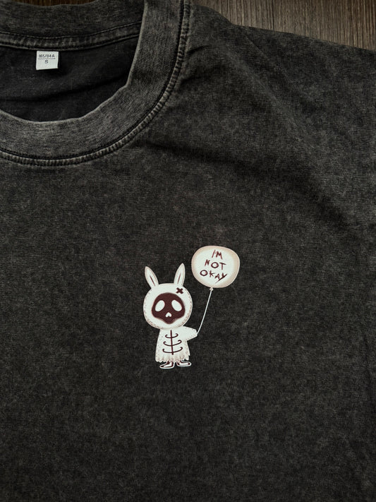 A close-up of a dark gray mineral-wash cotton t-shirt featuring a small graphic of a white bunny skeleton holding a balloon that says 'I'M NOT OKAY' in My Chemical Romance “The Black Parade” font