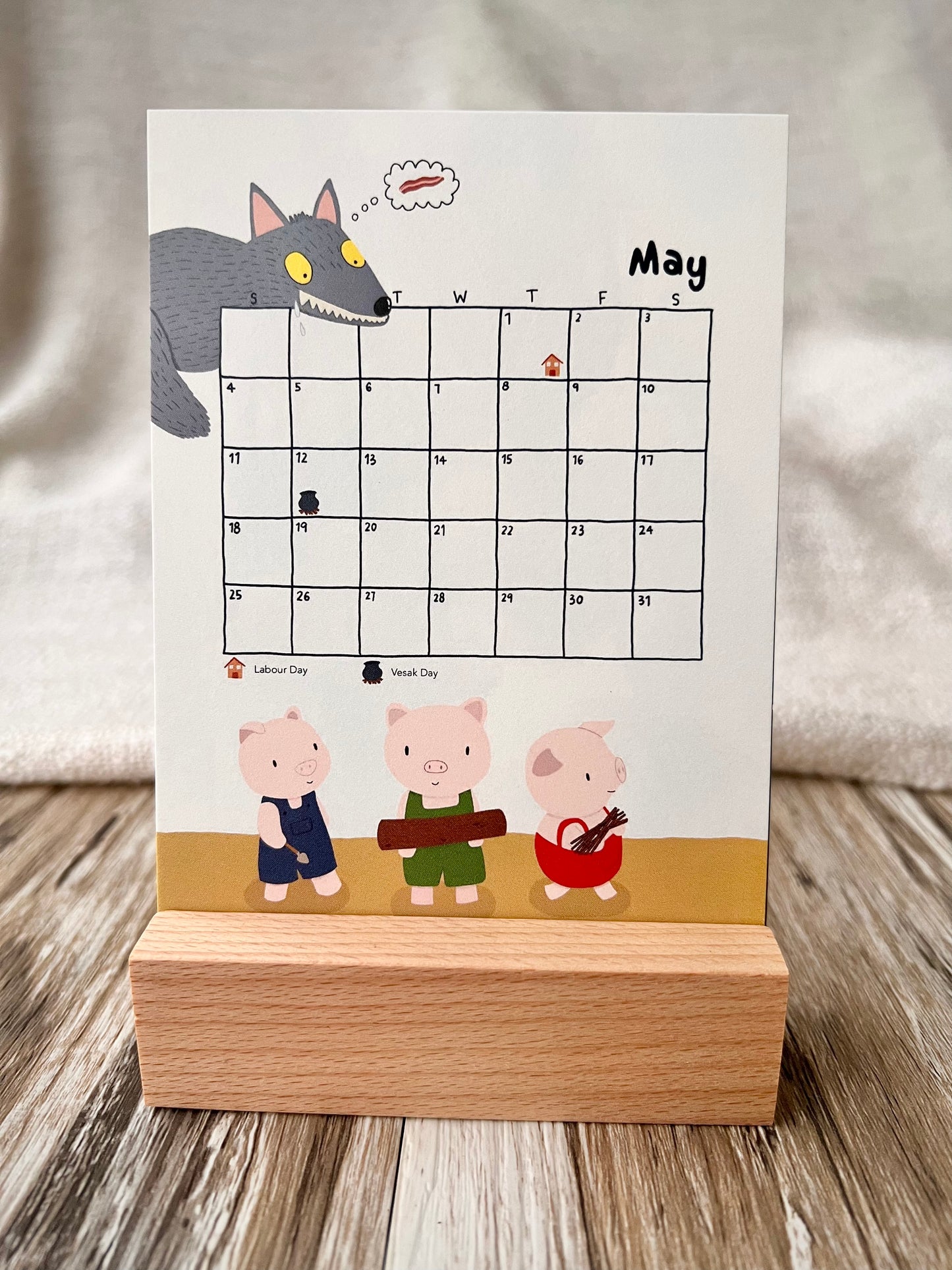 May 2025 calendar inserted in the wooden stand on a desk. The illustration of the May calendar is the tory of "Three Little Pigs", the Big Bad Wolf is watching Three Little Pigs and drooling. The public holiday is marked. 