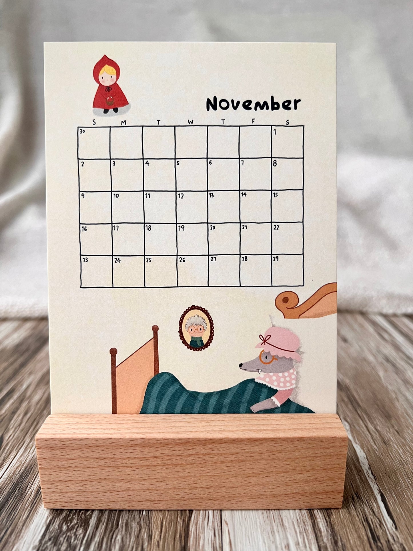 November 2025 calendar inserted in the wooden stand on a desk. The illustration of the November calendar is the story of "The Red Riding Hood". The Big Bad Wolf is lying on the bed, dressing in grandma's clothes. Red Riding Hood is holding her basket, going to visit her grandma