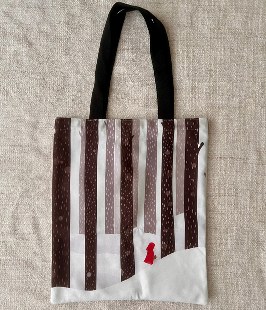 The image shows a cotton tote bag with illustration of red riding hood. The illustrations shows a tiny red riding hood, with her tiny basket, walking in the snow, among the tall trees. The colour makes red riding hood stood out perfectly. 