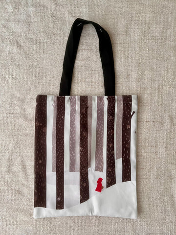 The image shows a cotton tote bag with illustration of red riding hood. The illustrations shows a tiny red riding hood, with her tiny basket, walking in the snow, among the tall trees. The colour makes red riding hood stood out perfectly. 