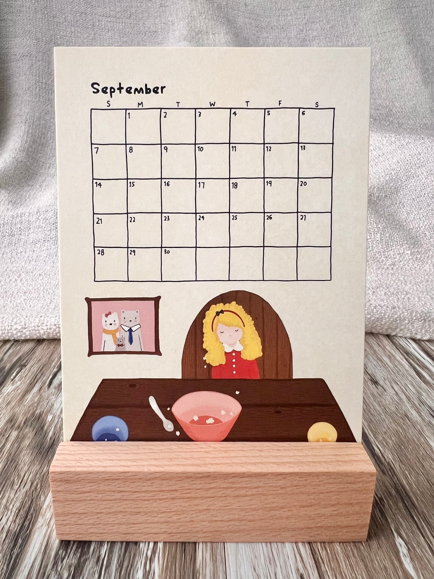 September 2025 calendar inserted in the wooden stand on a desk. The illustration of the September calendar is the story of "Goldilocks and the Three Bears", Goldilocks sat at the dining table, finished all the food. While there was a family portrait of the three bears at the background.