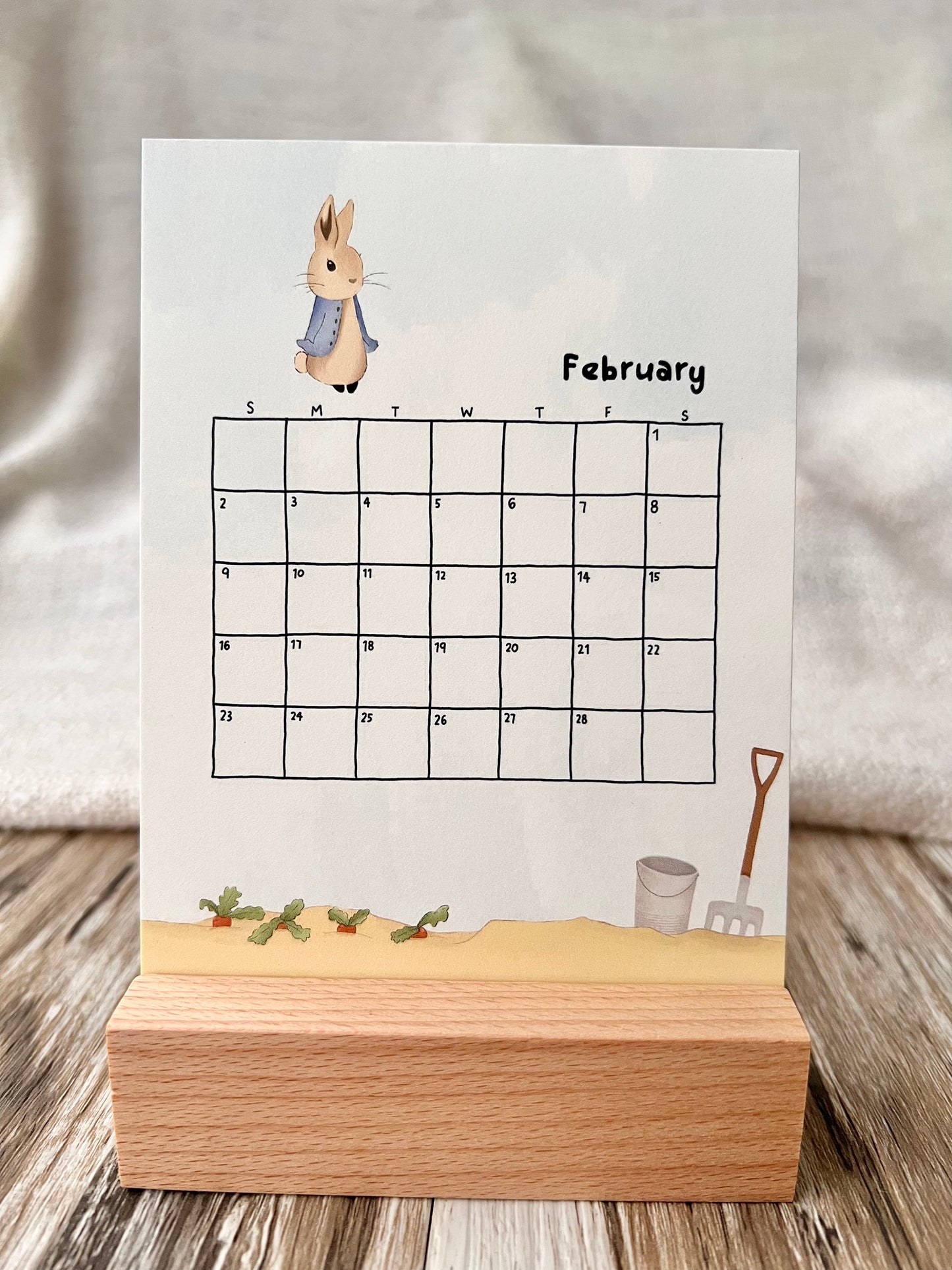 February 2025 calendar inserted in the wooden stand on a desk. The illustration of the February calendar is Peter Rabbit and the farm